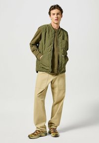 Gilet imbottito verde oliva con tasca frontale, abbinato a un top a maniche lunghe coordinato e pantaloni beige chiaro, e scarpe da trekking marroni.