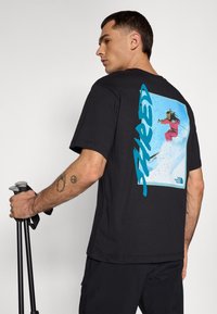 Black cotton t-shirt featuring a colored graphic of a skier on the back, with "SHRED" printed in blue on the side. The model holds trekking poles.