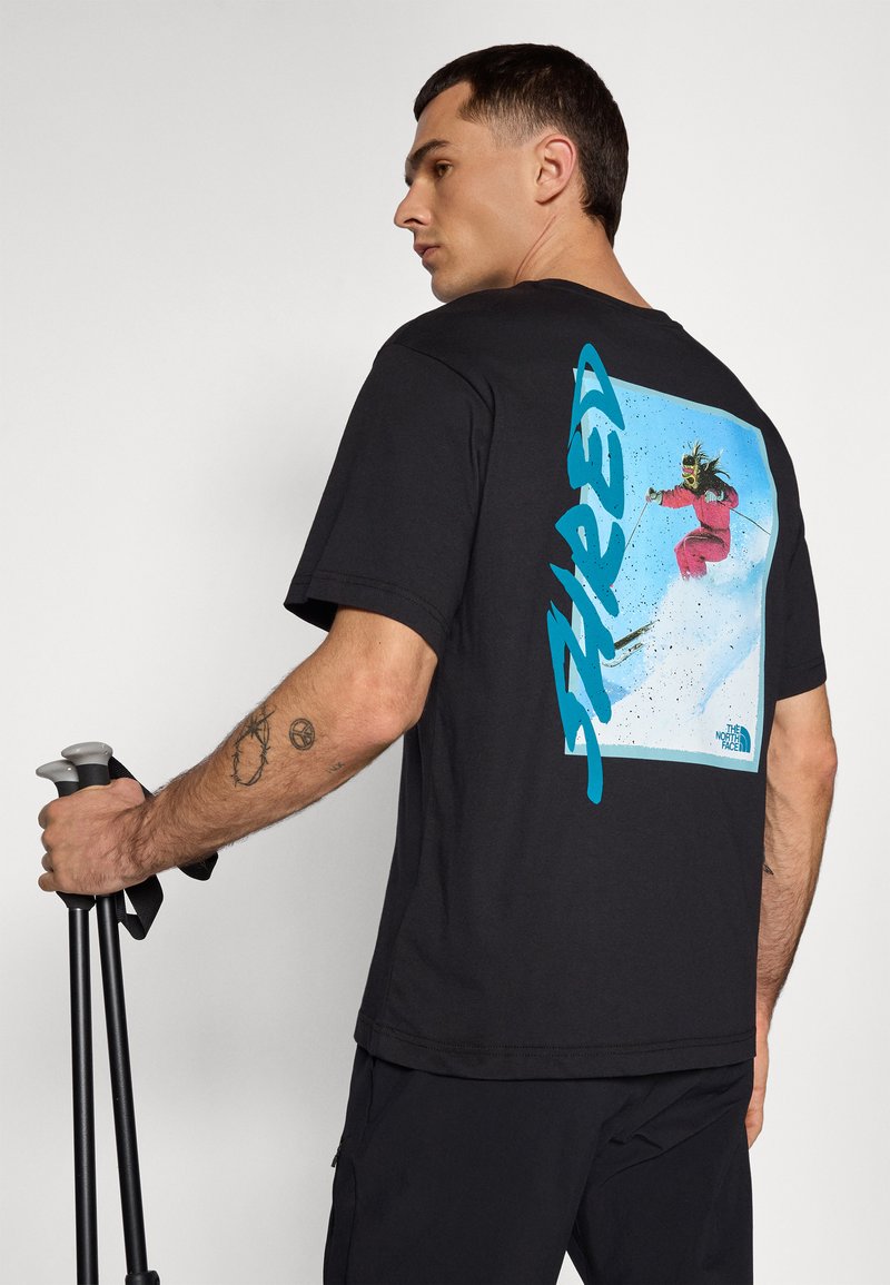 T-shirt en coton noir avec un graphique coloré d'un skieur au dos, avec "SHRED" imprimé en bleu sur le côté. Le modèle tient des bâtons de trekking.