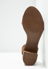 Semelle marron d’une sandale à talon pour femme avec adhérence texturée et talon bloc, reposant contre un angle de mur et de sol blancs.