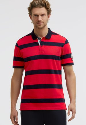 Mann mit lockigem Haar, der ein rotes Poloshirt mit breiten navyblauen horizontalen Streifen und schwarze Hosen trägt, steht vor einem schlichten weißen Hintergrund.