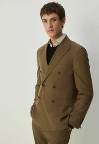 Brūns divkrūšu uzvalks ar melniem pogām, šaurs piegriezums, kombinēts ar melnu džemperi un gaišu krekla, uz neitrālas fona.