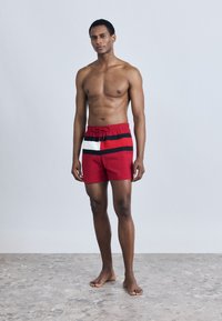 Κόκκινα μαγιό σορτς με μαύρη και λευκή ριγέ λεπτομέρεια, ελαστική μέση με κορδόνι και ελαφρύ ύφασμα.