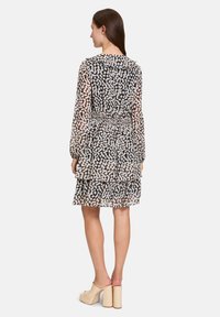Zwarte en beige gemêleerde transparante jurk met een gerimpelde zoom, smock taille en lange mouwen; model draagt beige blokhak muiltjes.