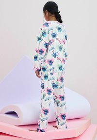 Valkoiset fleece-pyjamat, joissa on monivärisiä Stitch-hahmotulosteita, löysä istuvuus ja joustavat hihansuut sekä yläosassa että housuissa.