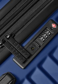 Gros plan sur une valise bleue avec des doubles fermetures éclair noires, verrouillée par un cadenas à combinaison TSA, affichant le nom de la marque Redolz.