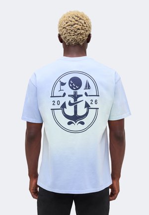 Person med kort krøllet blondt hår iført en hvid skjorte med en stor navy anker- og nautiske symboler grafik på ryggen.