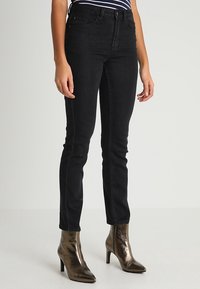 Jean skinny noir taille haute, avec un design classique à cinq poches et des coutures discrètes. Des bottines à talons brillantes complètent le look.