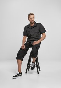Juodas trumpomis rankovėmis marškinėliai su dviem krūtinės kišenėmis, derinami su juodais kargo šortais ir juodais sportiniais bateliais, sėdintis ant juodos taburetės.