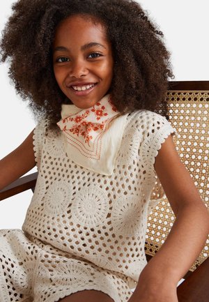 Tenue en crochet couleur crème avec des motifs circulaires ; associée à une écharpe ivoire ornée de détails floraux orange. Fond avec une chaise en bois naturel.