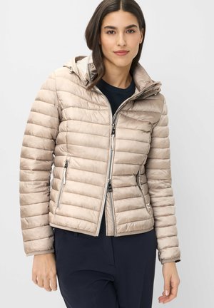 Frau trägt eine beige gesteppte Jacke mit Reißverschluss und Kapuze, kombiniert mit dunklen Hosen, stehend vor einem einfarbigen hellen Hintergrund.