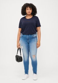 A woman with curly hair stands in front of a plain background, wearing a navy t-shirt, ripped blue jeans, and holding a small black bag.