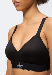 Γυναίκα που φοράει μαύρο bralette Calvin Klein με φαρδιές τιράντες και ελαστική ταινία με το σήμα κάτω από το στήθος, εμφανιζόμενη από το στήθος και πάνω.