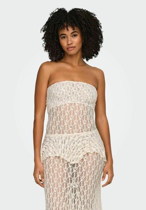 Kvinde med krøllet hår iført en stropløs, gennemsigtig hvid blondekjole med en flæsepeplumdetalje mod en ensartet baggrund.