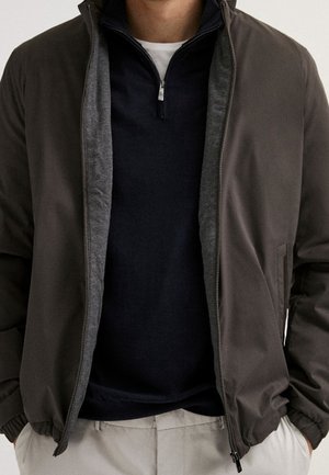 Homme portant une veste zippée marron foncé par-dessus un pull à fermeture éclair noire au col montant et un pantalon beige, les mains dans les poches de la veste, sur un fond uni.