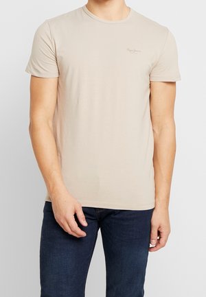 T-shirt beige à manches courtes col rond avec un petit logo sur la poitrine gauche, porté par une personne avec un jean bleu foncé sur un fond uni.