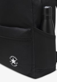 Mustast vastupidavast kangast koolikott, millel on külgmine võrktasku veepudeli jaoks, tõmblukuga sektsioon ja valged logodetailid.