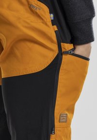 Gelbe und schwarze Outdoor-Hosen verfügen über Reißverschlusstaschen, flache Nähte und ein dreieckiges Logo-Patch. Das Material scheint leicht und langlebig zu sein.