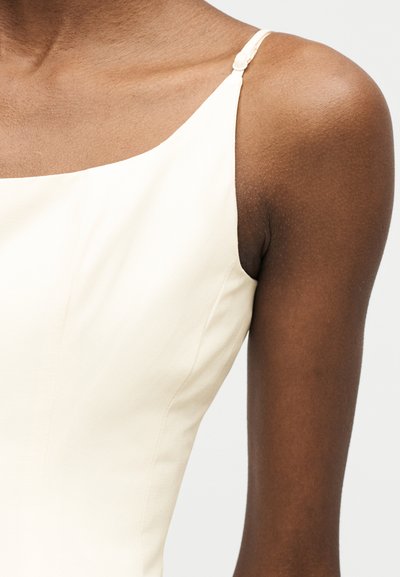 Άτομο με σκουρόχρωμη επιδερμίδα που φοράει αμάνικο, εκρού τοπ με λεπτό ρυθμιζόμενο λουράκι στον ώμο, σε απλό φόντο.