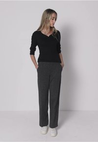 Schwarzes Langarmshirt mit V-Ausschnitt, kombiniert mit grauen, weit geschnittenen Hosen aus weichem Material. Das Model steht mit den Händen in den Taschen und trägt weiße Sneaker.