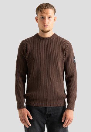 Homme portant un pull marron côtelé à manches longues et un pantalon noir, debout devant un fond gris clair uni.