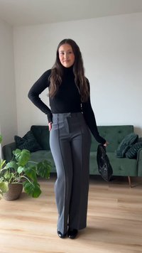 Γυναίκα με μακριά μαλλιά, ντυμένη με μαύρο ζιβάγκο, γκρι παντελόνι φαρδύ, κρατώντας μια μαύρη τσάντα, στέκεται σε δωμάτιο με πράσινο καναπέ και φυτά.