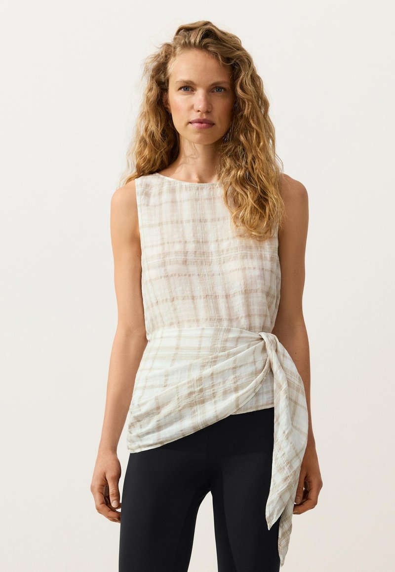 Femme aux cheveux blonds bouclés porte un haut beige à carreaux sans manches avec un détail noué sur le côté et un pantalon noir, debout devant un fond uni.