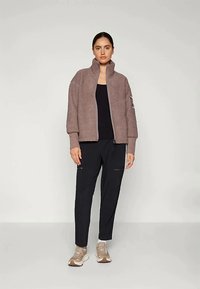 Γυναίκα που στέκεται φορώντας ένα ταμπά φούτερ με φερμουάρ, μαύρη μπλούζα, μαύρο παντελόνι και μπεζ αθλητικά παπούτσια, με φόντο απαλό ανοιχτό χρώμα.