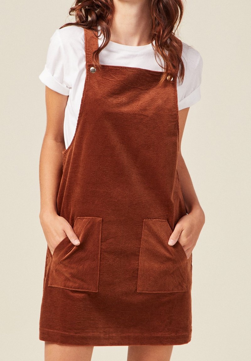 Femme portant une robe salopette en velours côtelé marron avec des poches avant sur un t-shirt blanc à manches courtes, mains dans les poches, sur un fond uni.