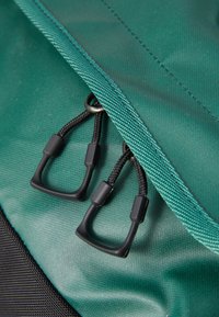 Green fabric backpack close-up, featuring a textured surface, black hardware, and cord pulls for secure closure.