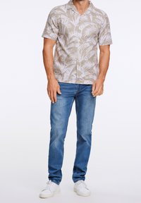 Chemise beige à manches courtes avec un motif de feuilles de palmier blanches, associée à un jean bleu et des baskets blanches, posant devant un fond clair.