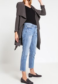 Grau drapierter Cardigan über einem schwarzen Oberteil, kombiniert mit hellblauen, gekürzten Jeans. Dunkle Slippers runden den Look ab.