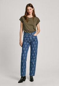 Tmavozelené tričko s krátkym rukávom skombinované s džínsami s vysokým pásom so vzorom v modrej farbe, ktoré majú kvetinové a geometrické vzory; nosené s čiernymi členkovými topánkami.