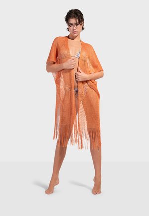 Kvinde iført en orange net-frynset cover-up over en blomsterbikini, stående barfodet mod en ensartet lys baggrund.