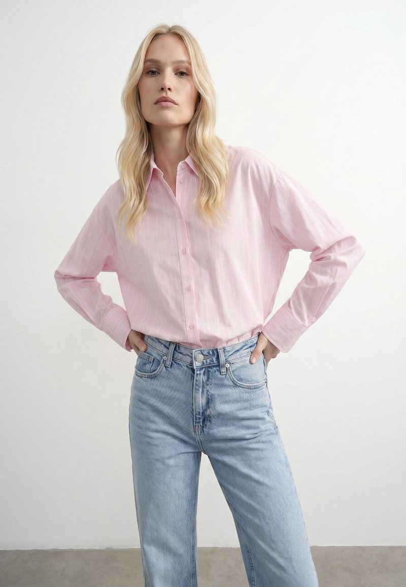 Femme blonde debout, les mains sur les hanches, portant une chemise rose clair boutonnée et un jean taille haute bleu clair, sur un fond blanc uni.