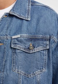 Denim jacket featuring a light blue wash, a classic collar, chest pocket with branded button, and visible stitching details.