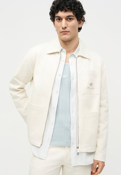 Νέος άντρας με σγουρά σκούρα μαλλιά που φορά στρωμένο ανοιχτόχρωμο σακάκι, πουκάμισο με κουμπιά και υφασμάτινο πλεκτό γιλέκο μπροστά σε απλό φόντο.