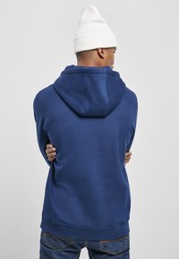 Felpa con cappuccio di un blu navy, realizzata in un morbido misto cotone, presenta una tasca a marsupio, polsini a costine e una vestibilità rilassata. Indossata con un berretto bianco.