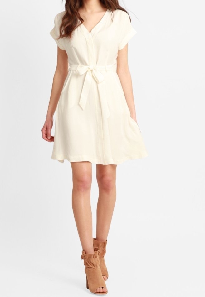 Robe enveloppante couleur crème avec des manches courtes, une ceinture à nouer à la taille et des poches latérales, fabriquée en tissu léger. Portée avec des bottines beiges.