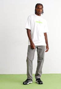 Hvid t-shirt med korte ærmer, der har et farverigt grafisk design, parret med lysegrå bukser og sorte sneakers med hvide detaljer.