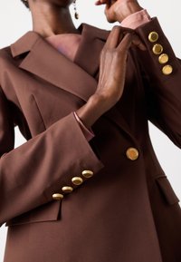 Brun skreddersydd blazer med dobbeltspent design, gullknappdetaljer og en glatt tekstur, med en lett glans og strukturerte skuldre.