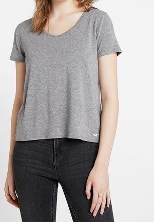Camiseta gris de manga corta con cuello en V, hecha de una tela suave, que presenta un ajuste ligeramente holgado y un pequeño logo blanco en el dobladillo inferior.
