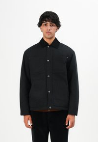 Young man with curly hair wearing a black button-up jacket with four front pockets, standing against a white background.
