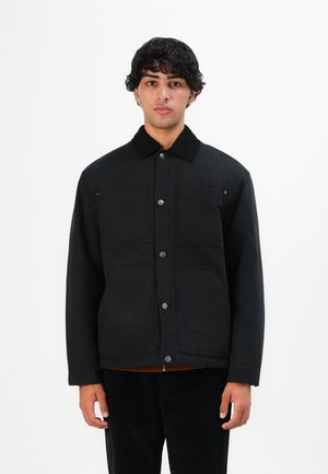 Young man with curly hair wearing a black button-up jacket with four front pockets, standing against a white background.