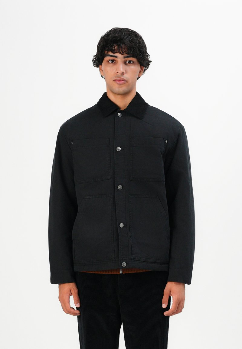 Young man with curly hair wearing a black button-up jacket with four front pockets, standing against a white background.