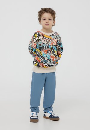 Felpa colorata con stampe grafiche, polsini beige, abbinata a pantaloni azzurri chiari e scarpe da ginnastica sportive con accenti bianchi e navy.