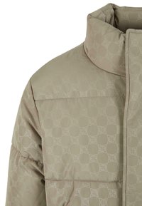 Beige pufferjakke med høj krave, der har et subtilt geometrisk mønster. Den er lavet af quiltet materiale og har en glat tekstur.