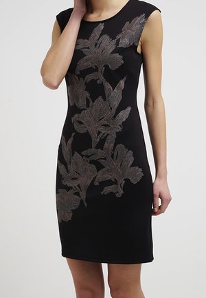 Ärmelloses schwarzes Kleid mit großen metallischen Blumenmustern von der Schulter bis zum Saum, getragen von einer Person mit der Hand nahe am Gesicht vor einfarbigem Hintergrund.