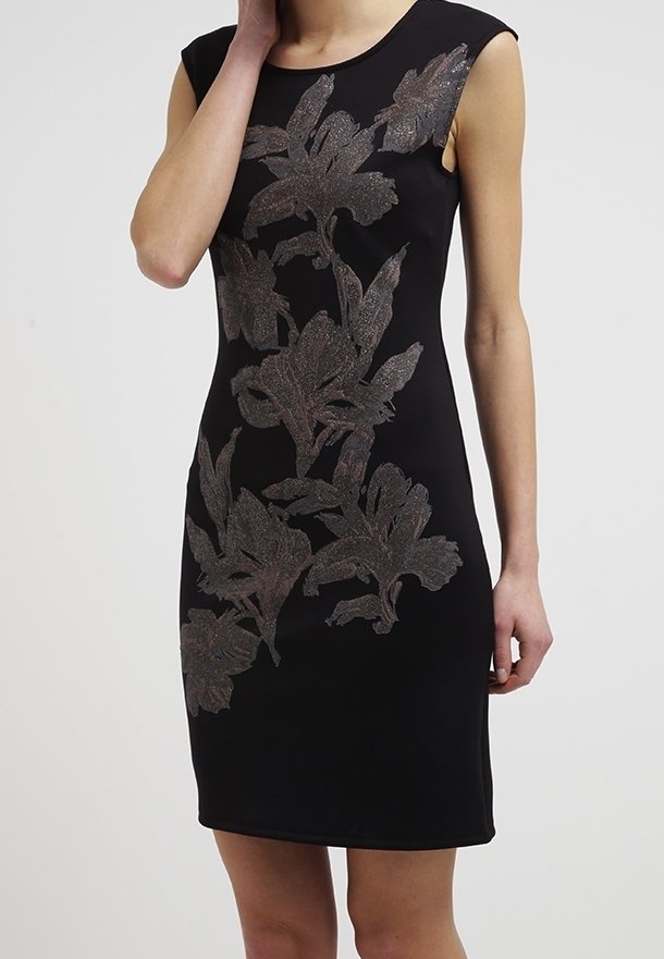 Robe noire sans manches avec de grands motifs floraux métalliques de l'épaule à l'ourlet, portée par une personne avec la main près du visage sur un fond uni.