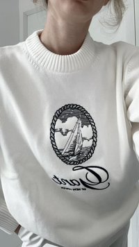 Suéter de punto blanco con un parche bordado redondo que presenta un velero rodeado de nubes, delineado con un diseño de cuerda. Texto negro debajo del parche.